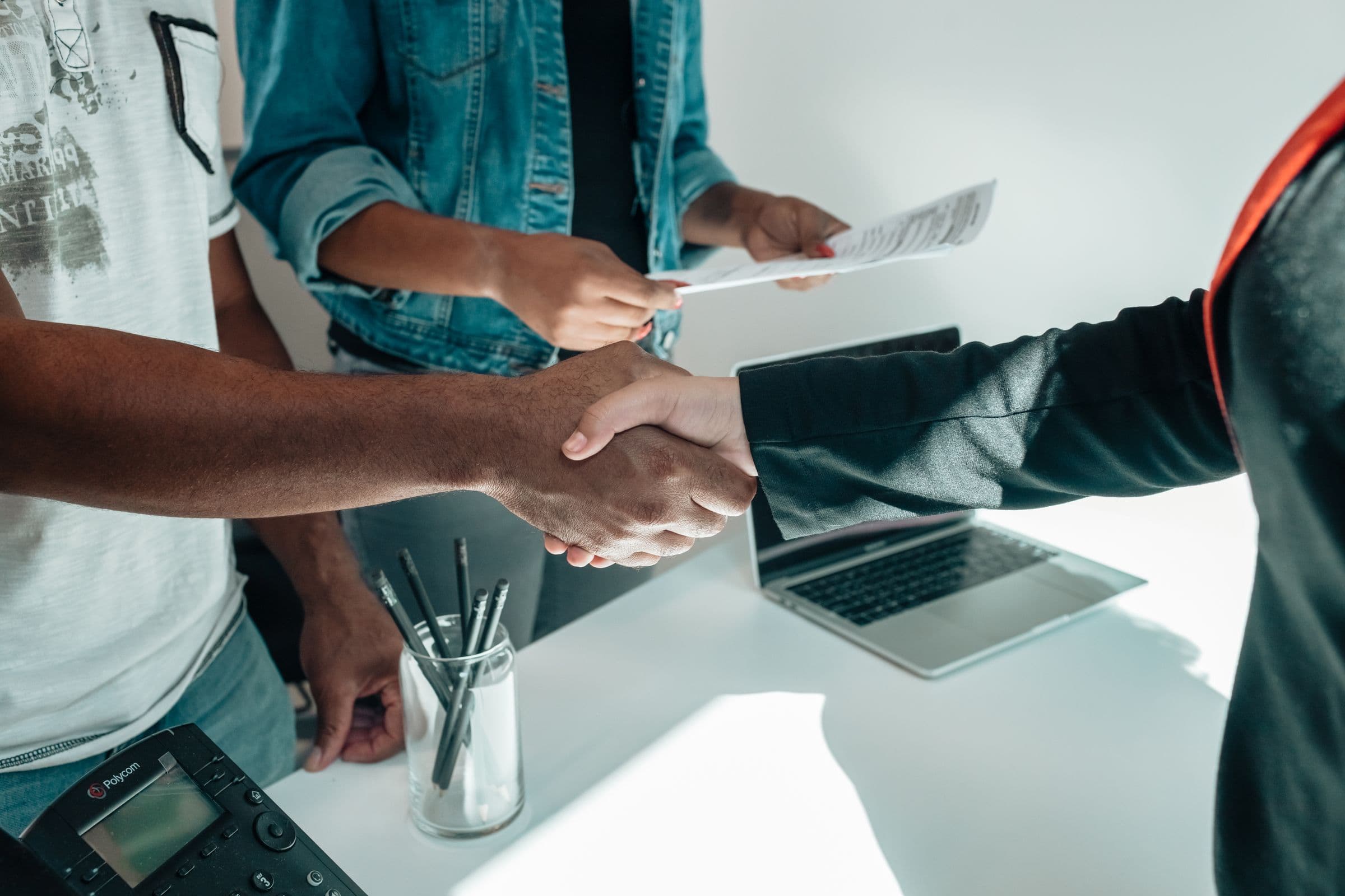 Business partners shaking hands across a sunlit desk with a laptop and documents