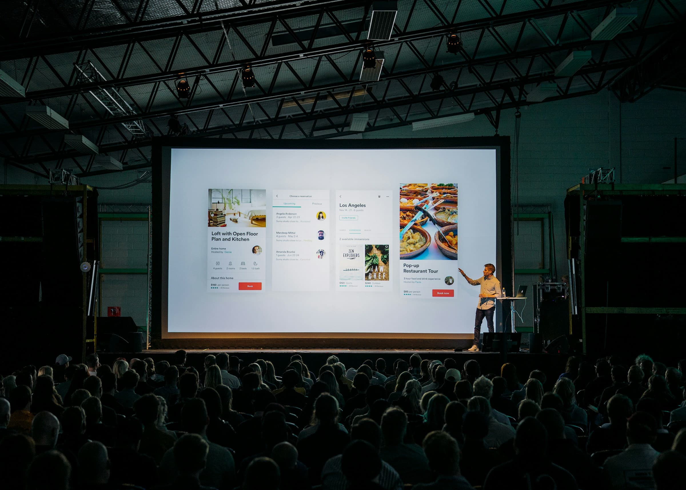 Wide view of a conference presentation with a large screen, speaker, and seated audience